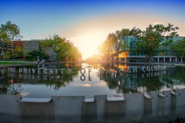 Under a clear blue sky, the sun sets behind a campus water fountain which the words "The University of British Columbia" float on top of.